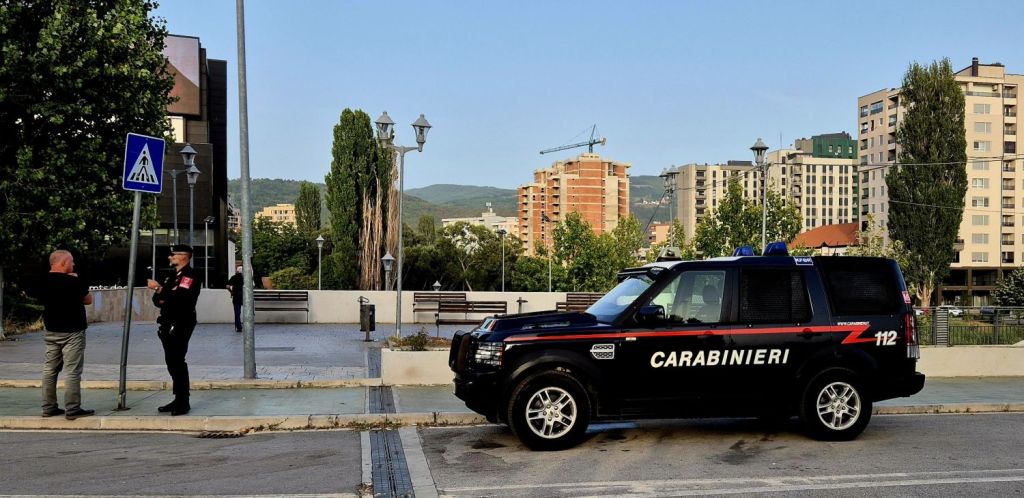 A Carabinieri police vehicle parked on the street, with an officer engaged in conversation with a person nearby. The background features buildings and trees.