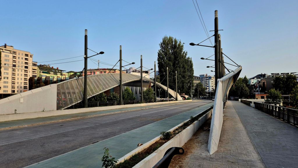 A modern bridge with a unique architectural design is shown spanning a wide road, flanked by tall trees and contemporary buildings under a clear blue sky.
