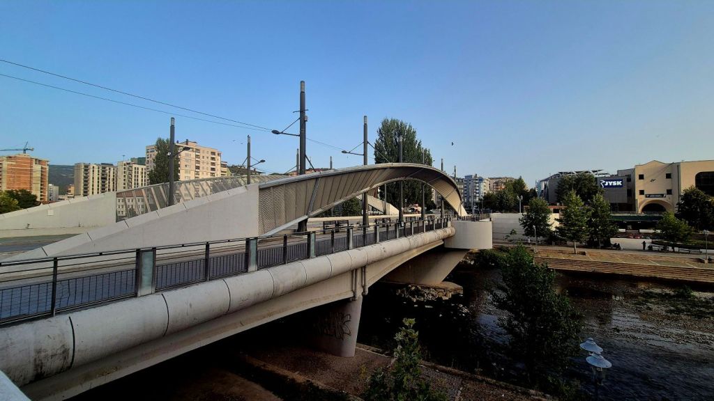 A modern arched bridge spanning a river, with urban buildings in the background and clear blue skies.