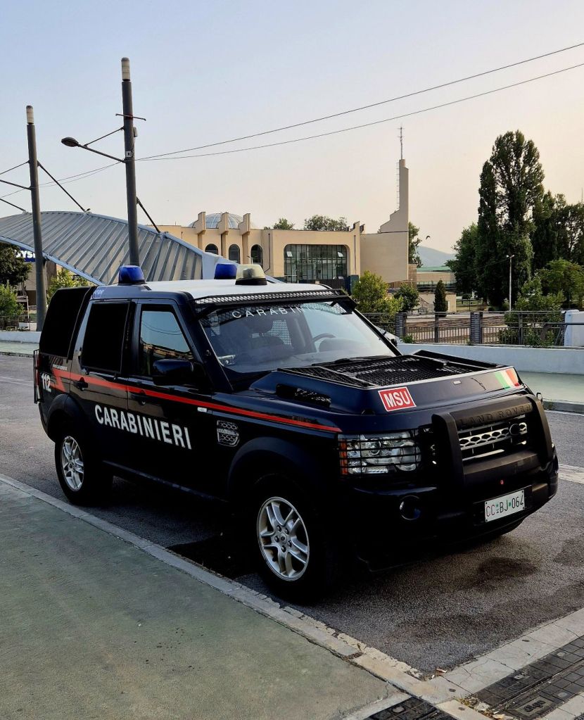 A black Carabinieri vehicle parked on a street, with blue emergency lights on top and a sign indicating 'MSU'. In the background, a modern building and trees are visible.