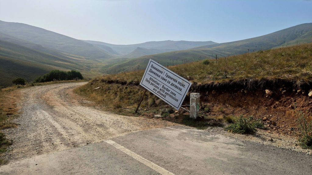 A winding dirt road leads into a mountainous landscape with rolling hills in the background. A signpost is prominently displayed on the roadside, warning of a restricted border zone.