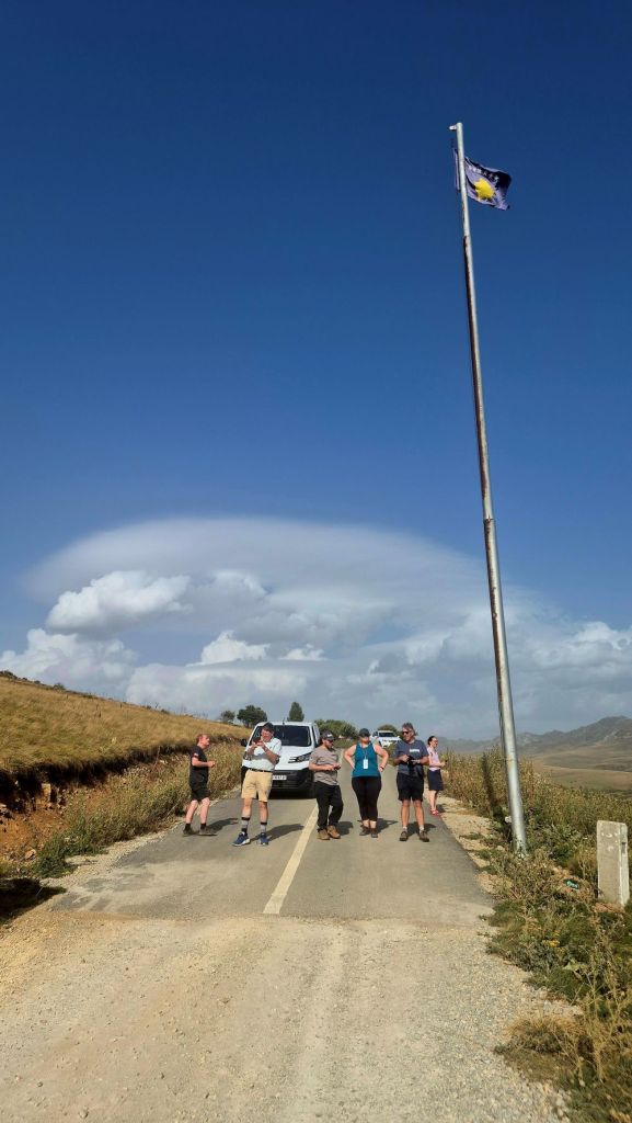 A group of five people standing on a rural road, with a car parked nearby and a flagpole displaying a flag under a clear blue sky.