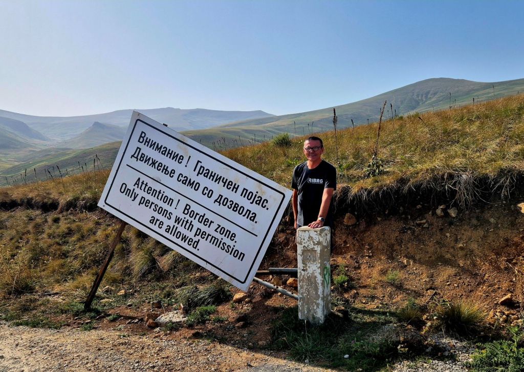 A person standing next to a large sign that reads 'Attention! Border zone: Only persons with permission are allowed.' in both Russian and English, set against a scenic mountainous landscape.