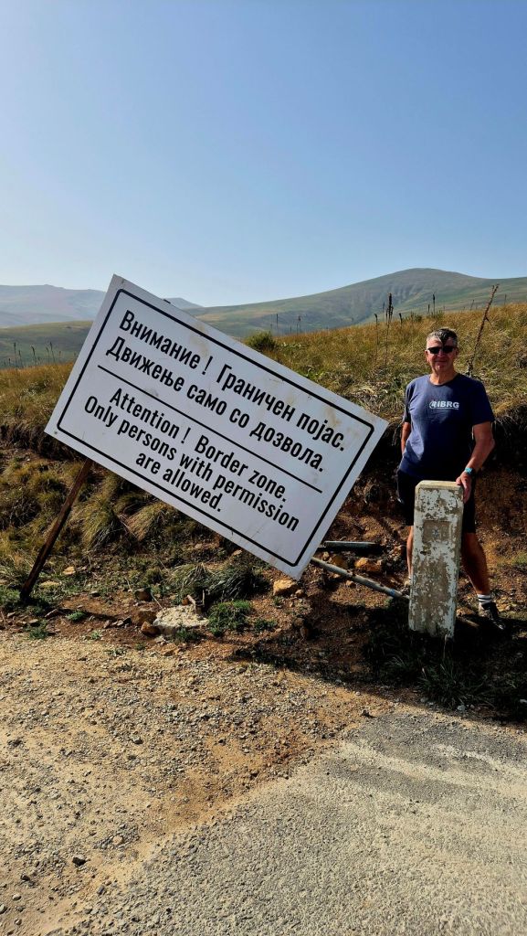 A person standing next to a warning sign stating 'Attention! Border zone. Only persons with permission are allowed.' The background features a hilly landscape under a clear blue sky.