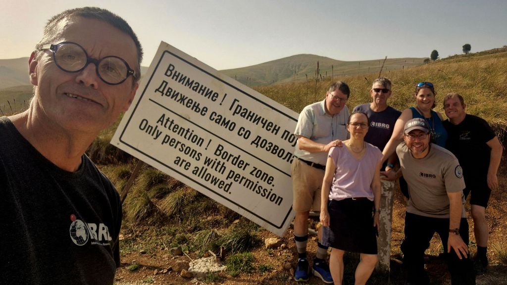 A group of seven people posed in front of a warning sign that reads 'Attention! Border zone. Only persons with permission are allowed.' The background features hilly terrain and grasslands.