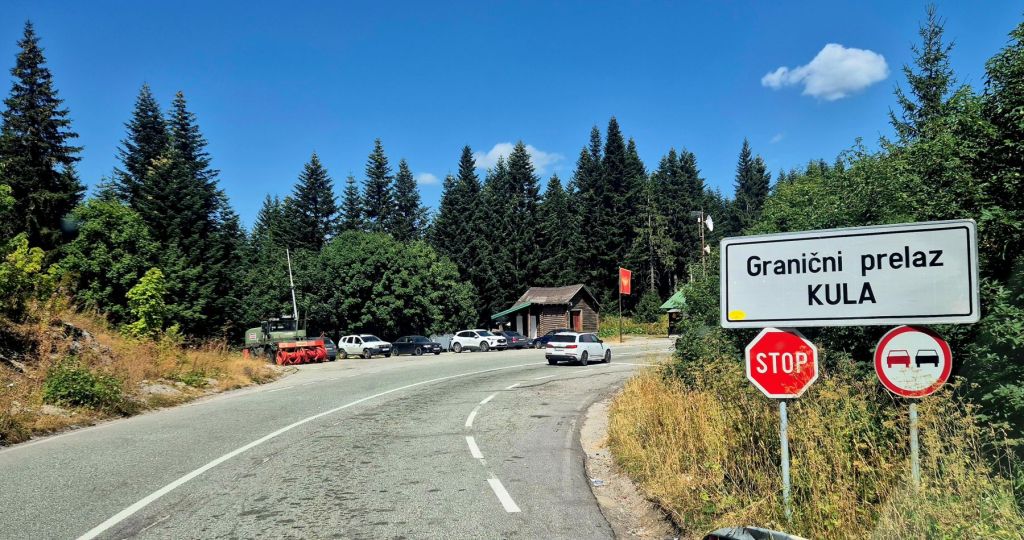 A roadside view featuring a border crossing sign for 'Granični prelaz KULA', surrounded by green trees and a clear blue sky. Several vehicles are parked nearby, and a stop sign is visible.