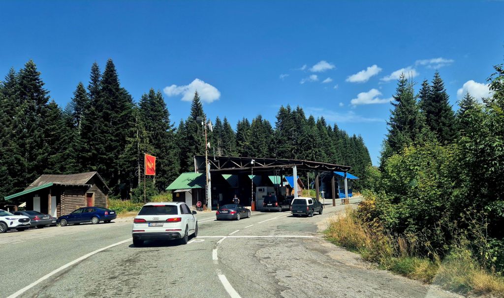 A view of a border checkpoint with vehicles waiting in line, surrounded by dense green forests and a clear blue sky.