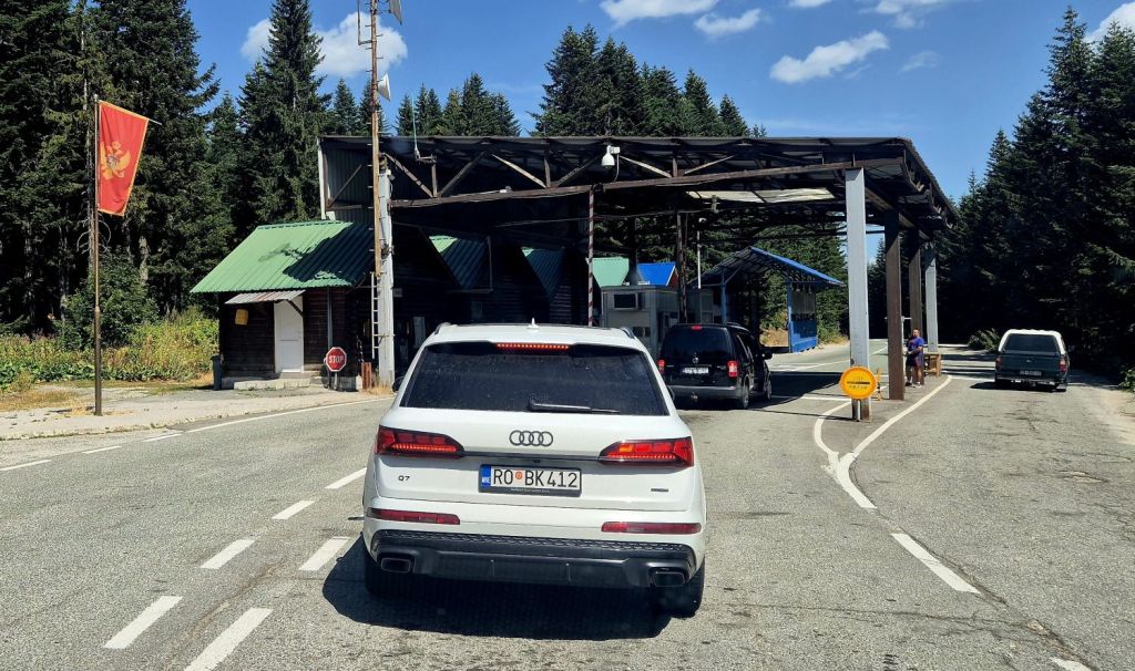 A white Audi SUV waiting in line at a border checkpoint surrounded by dense trees and a blue sky.