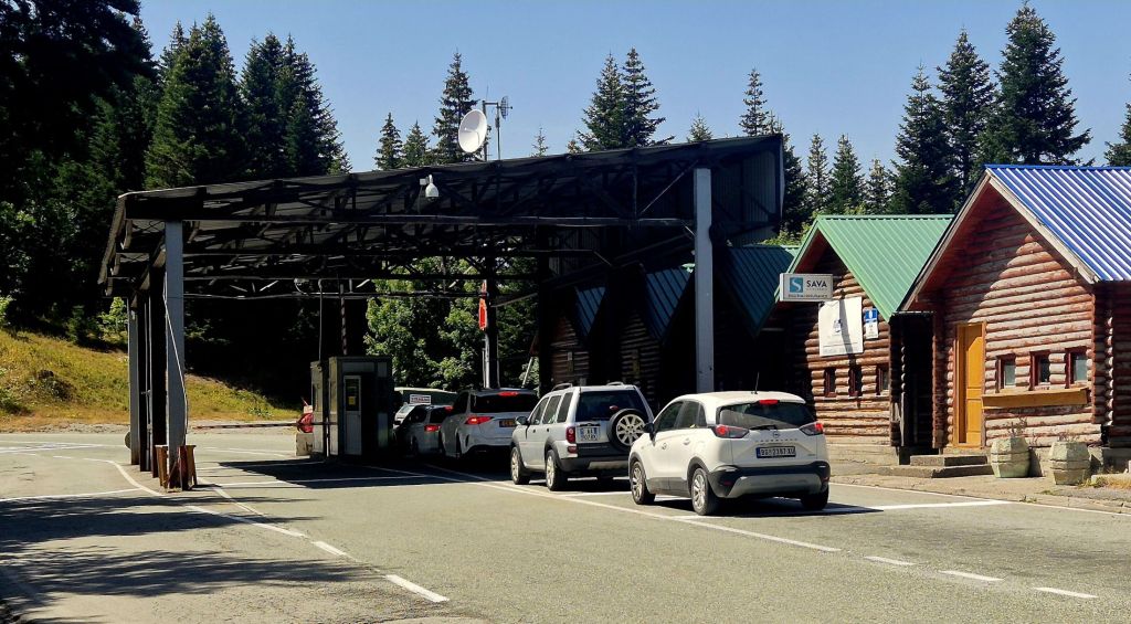 A vehicle checkpoint with a canopy, surrounded by trees and two wooden cabins, one displaying a sign. Cars are lined up at the toll booth.