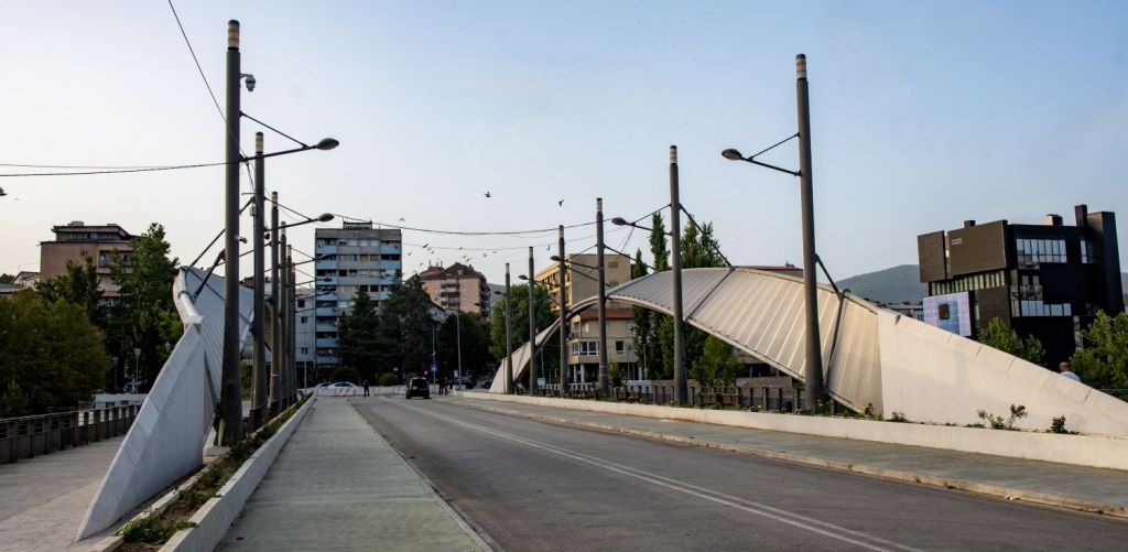 A modern bridge with a unique design spans a street, surrounded by buildings and trees under a clear sky.