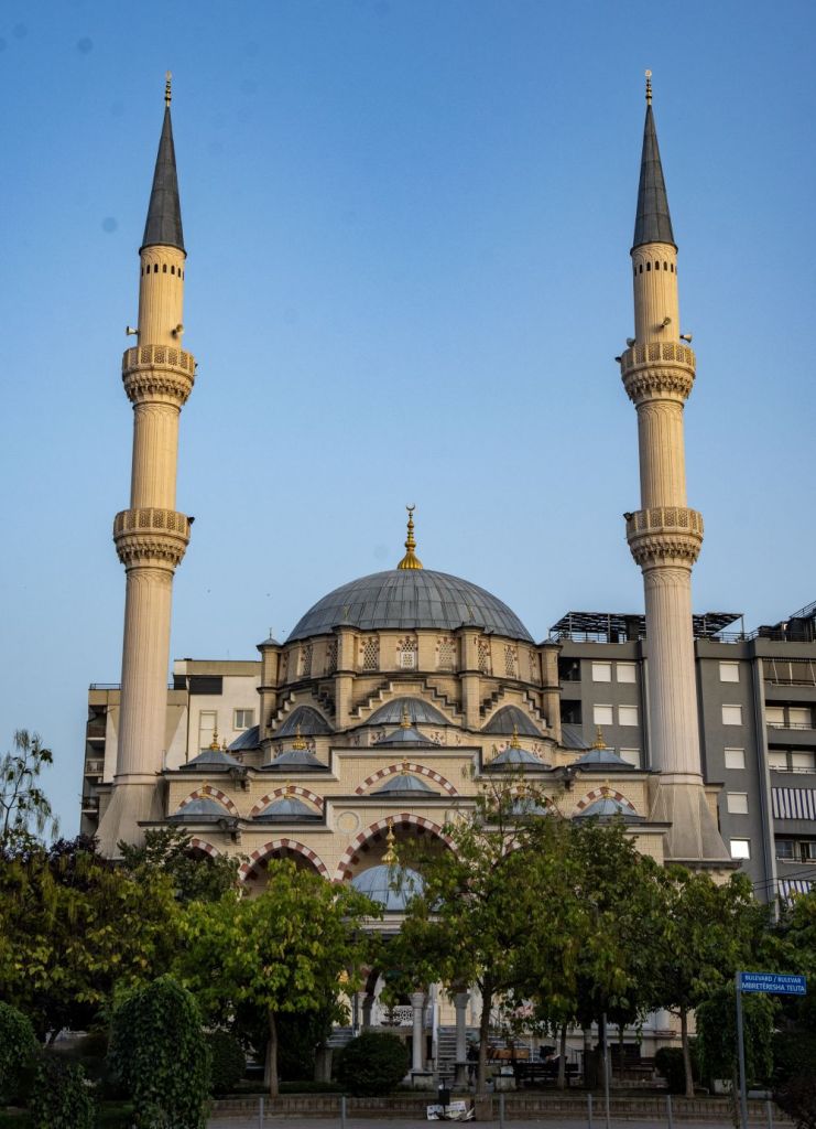 A mosque with two tall minarets and a large dome, surrounded by greenery and urban buildings under a clear blue sky.