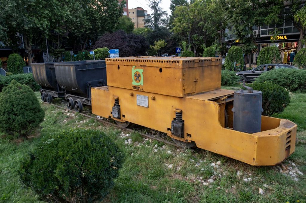 A yellow mining locomotive displayed in a park area, with two black coal wagons attached, surrounded by neatly trimmed bushes and greenery.