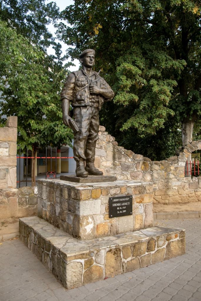 Bronze statue of a soldier standing on a stone pedestal, surrounded by trees, commemorating Shemsi Ahmeti, with an inscription detailing his dates of birth and death.