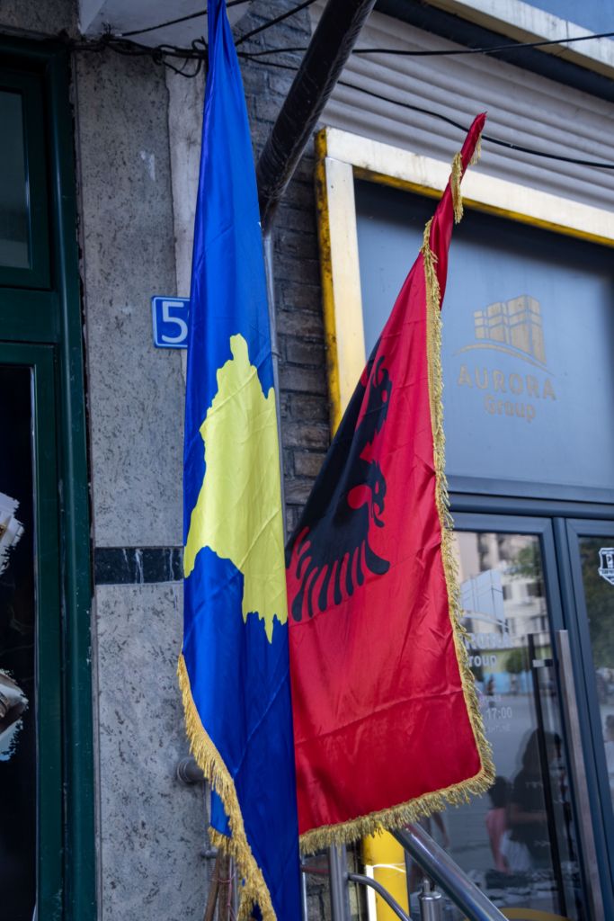 Two flags are displayed together: a blue flag with a yellow map of Kosovo and a red flag featuring a black double-headed eagle, symbolizing Albania.