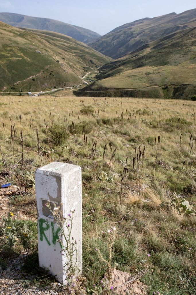 A concrete milestone marked with 'R' in a grassy landscape, overlooking a winding road in a valley surrounded by hills.