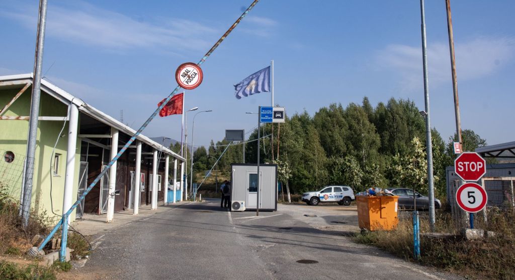 Border crossing entrance with flags, security booth, and stop sign in a rural area.