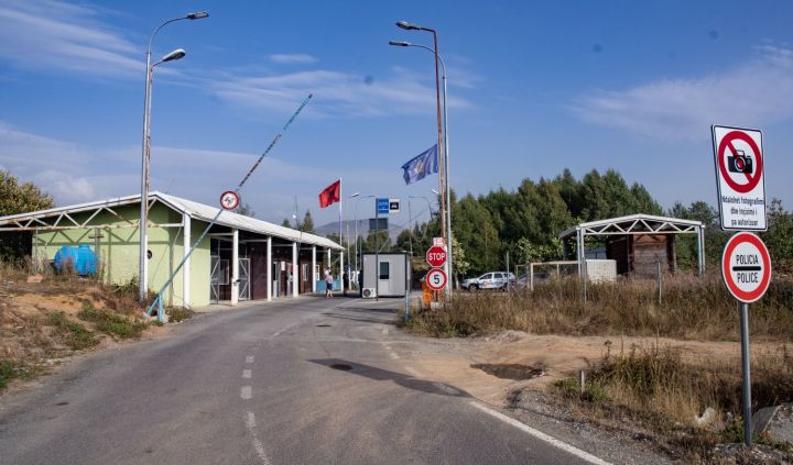 A road leading to a border crossing station, featuring flags, traffic signs, and a checkpoint structure surrounded by greenery.