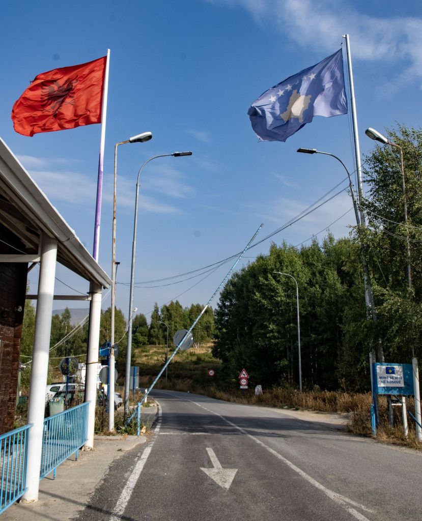Road entrance with Albanian and Kosovo flags waving in the background, surrounded by trees and highway signage.
