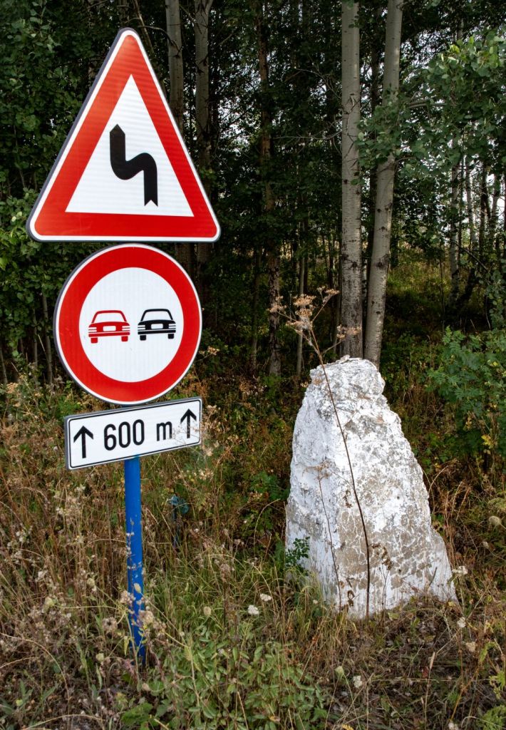 A road sign indicating a sharp turn ahead, accompanied by a no overtaking sign, with a distance marker of 600 meters. The signs are located next to a white rock and surrounded by greenery.