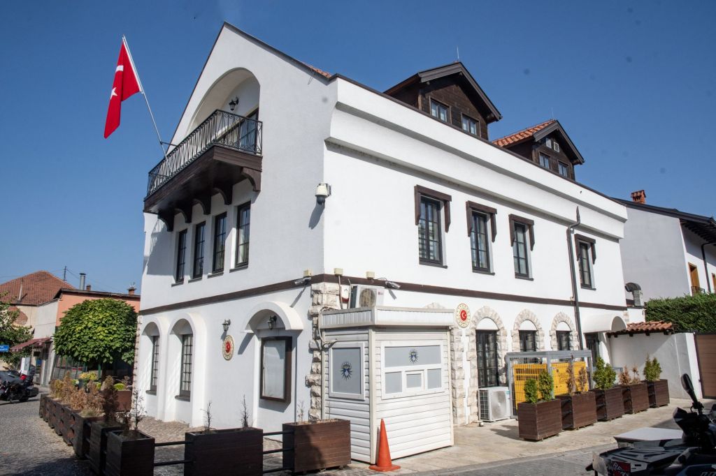 A white building with a decorative balcony and a red Turkish flag, surrounded by greenery and a cobblestone path.