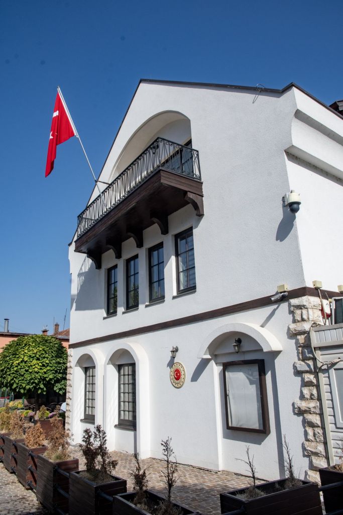 A white building with a balcony, featuring a Turkish flag and decorative elements, surrounded by green landscaping.