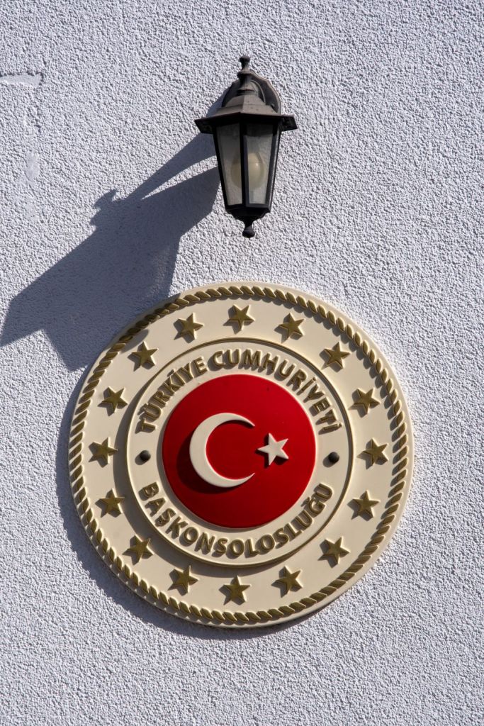 Emblem of the Turkish Republic Consulate, featuring a red circle with a white crescent and star, surrounded by gold stars and rope design, mounted on a white wall with a lantern above.