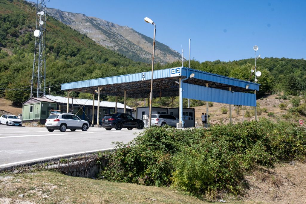 Border checkpoint with a blue roof, surrounded by vehicles and mountains in the background.