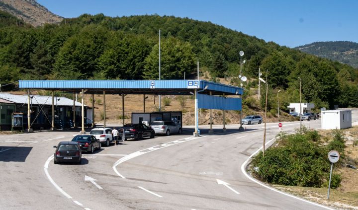 A border crossing checkpoint with several cars waiting in line, set against a backdrop of green hills and trees under a clear sky.