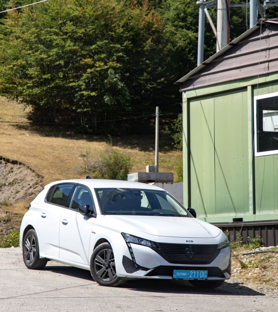 A white hatchback car parked near a green building, surrounded by trees and grass.