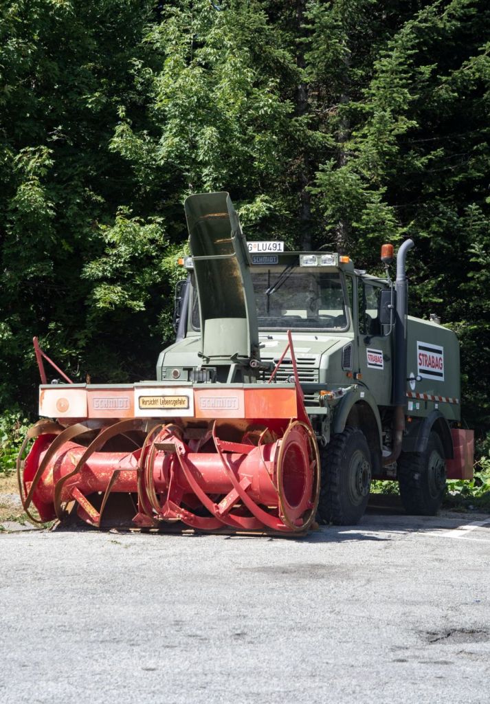 A snow plow truck with a large red snowblower attachment parked on a gravel road surrounded by green trees.