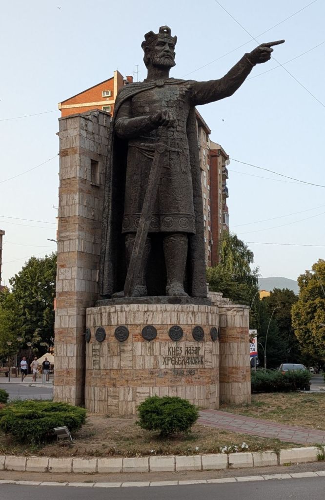 A large statue of a king pointing, made of dark stone, located in a public square. The statue stands atop a stone pedestal with inscriptions and symbols, surrounded by greenery and urban buildings.