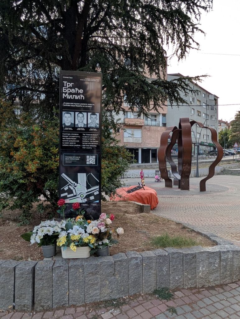 Memorial sign dedicated to the Brane Milic brothers with flowers in front, featuring details and images on a black informational panel surrounded by lush greenery and an abstract sculpture in the background.