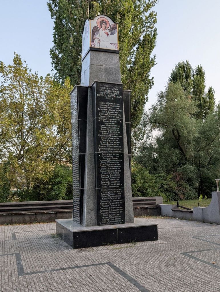 A tall memorial monument featuring an angelic figure at the top and black plaques listing names, surrounded by greenery and trees.