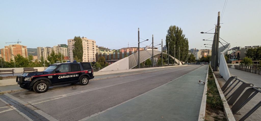 A Carabinieri police vehicle parked on a wide, empty road near a modern pedestrian bridge with urban buildings in the background.
