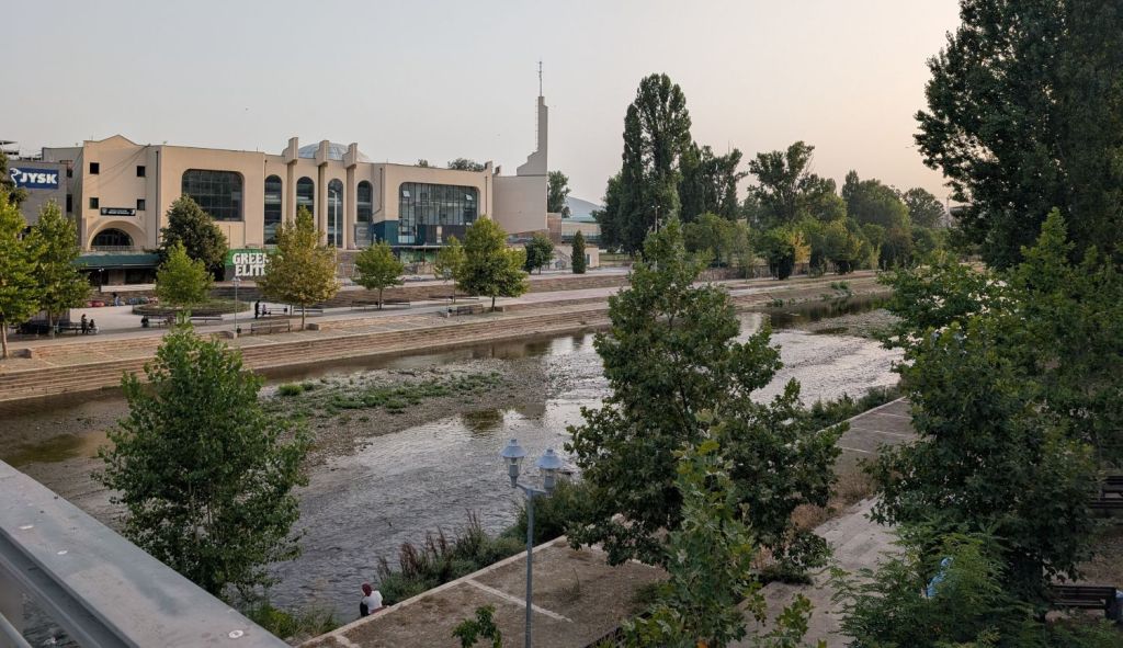 View of a riverbank with sparse water and greenery, featuring a modern building in the background.