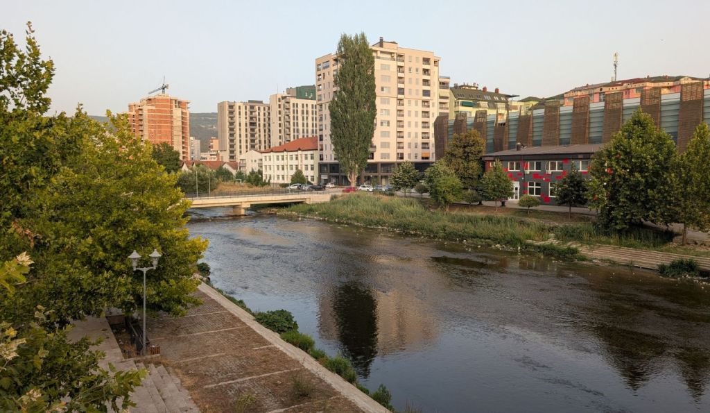 A riverside view showcasing modern buildings and greenery along the water, with a bridge crossing the river and urban architecture in the background.