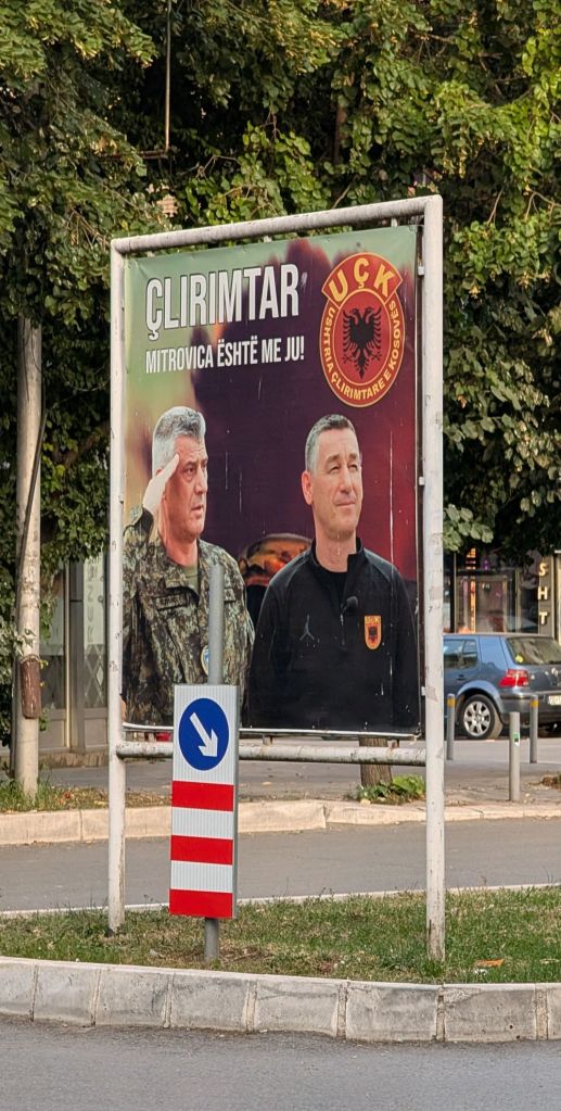 A billboard featuring two men, one in military uniform saluting, and the other in a black shirt, with the text 'ÇLIRIMTAR MITROVICA ËSHTË ME JU!' in Albanian.