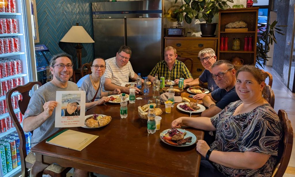 A group of seven people gathered around a table enjoying a meal, with various dishes including meat and bread on their plates. One person holds a sign that says 'Get Well Soon, Pete!' in front of them. The setting appears to be a cozy dining area with a soda vending machine in the background.