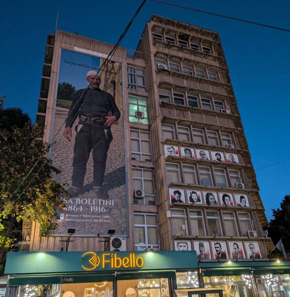 A large mural of a man in traditional attire on the side of a building, featuring a quote at the top. The building is multi-storied with several windows and air conditioning units, and a shop named 'Fibello' at the bottom.