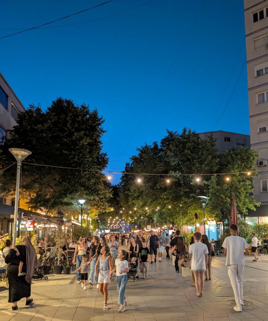 A bustling pedestrian street at dusk, filled with people walking, dining, and enjoying the ambiance under string lights and a blue sky.