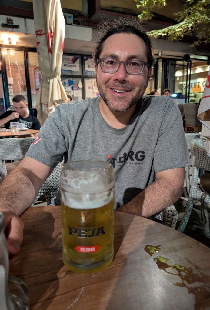 A man with glasses and long hair smiles while sitting at a table outdoors, holding a large glass of beer labeled 'Peja Pilsner'. In the background, another person can be seen at a nearby table.