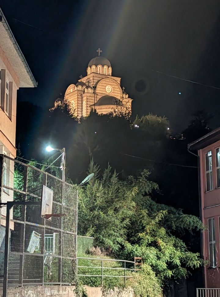 Night view of a church illuminated on a hill, surrounded by trees and buildings, with a basketball hoop in the foreground.