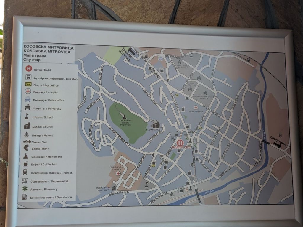 A city map of Kosovska Mitrovica displaying various landmarks, including hotels, bus stops, post offices, a hospital, police office, university, school, market, church, monument, coffee bars, train station, supermarket, pharmacy, and gas station.