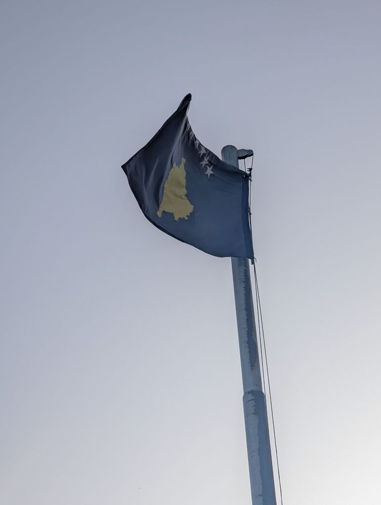 A flag featuring a yellow map silhouette against a dark background, flying on a flagpole with a clear sky in the background.