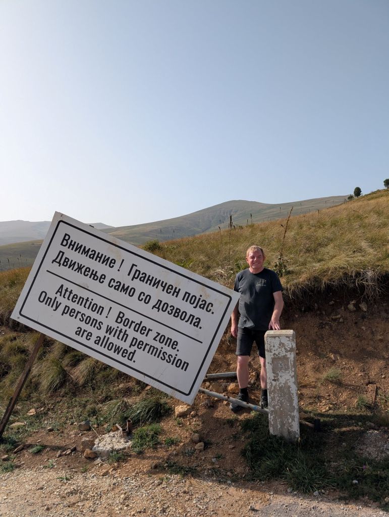 A man standing beside a large warning sign in a border area, with mountains in the background. The sign reads 'Attention! Border zone. Only persons with permission are allowed.'
