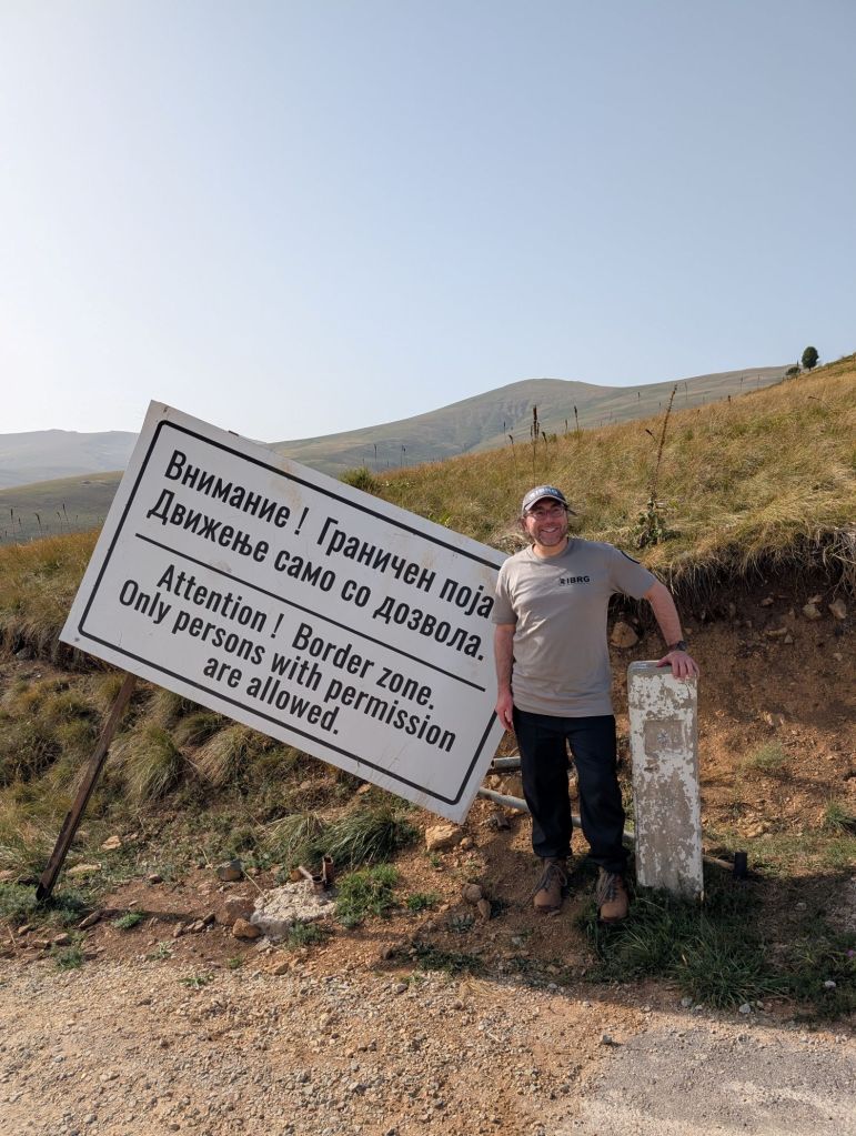 A man stands next to a large sign that reads 'Attention! Border zone. Only persons with permission are allowed.' The background features rolling hills and clear blue skies.