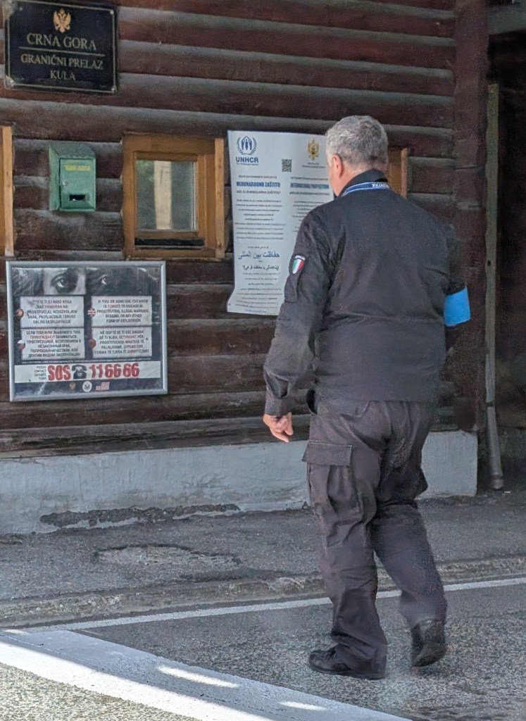 A person wearing a black uniform walks past a wooden building marked with signs indicating it is a border crossing in Montenegro, featuring various informational posters and a mailbox.